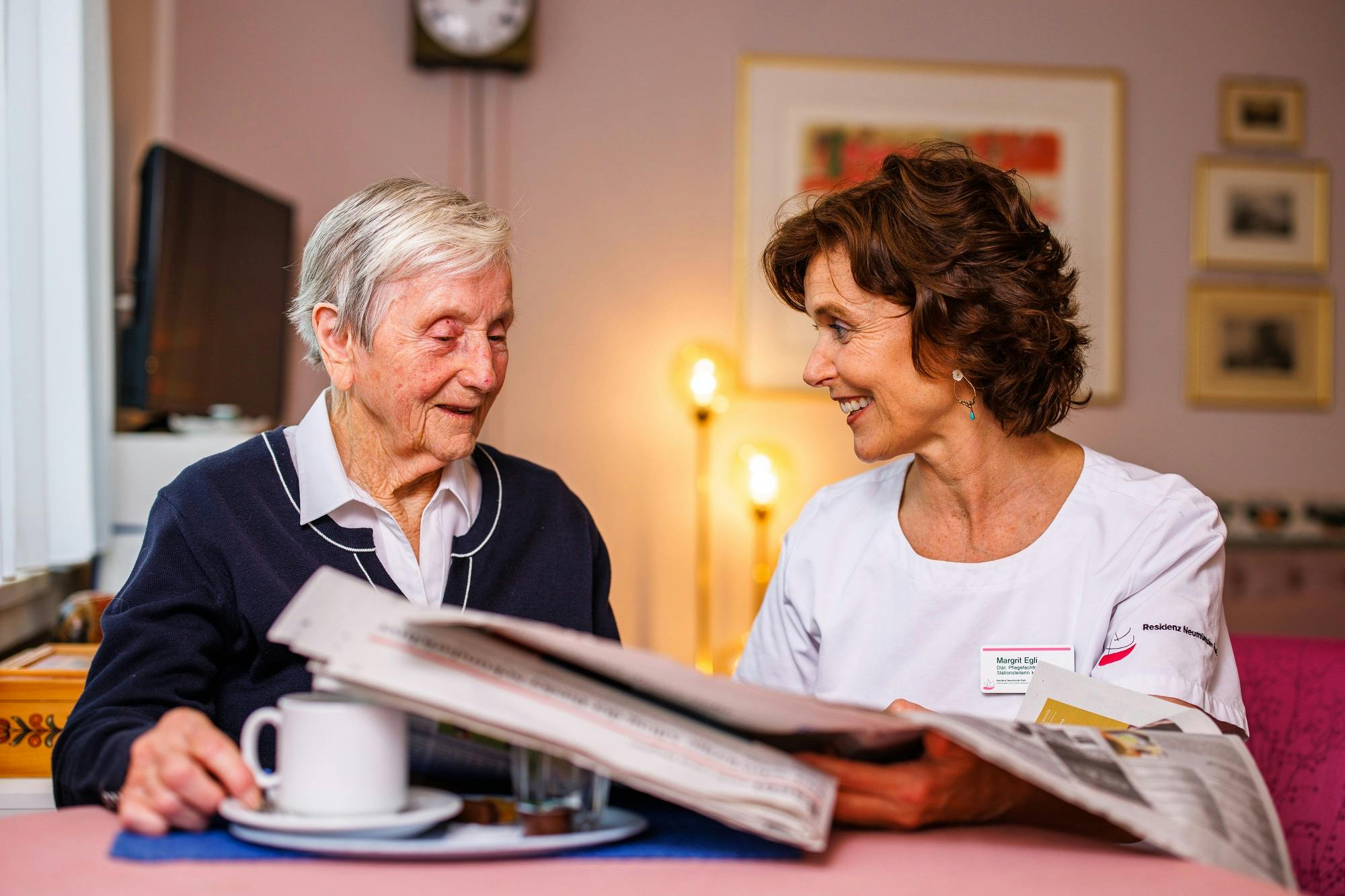 Elderly woman and carer smiling with a newspaper in a cosy room.