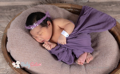 Neugeborenes schläft in einem Korb, umhüllt von einem lila Tuch und mit einem Stirnband.