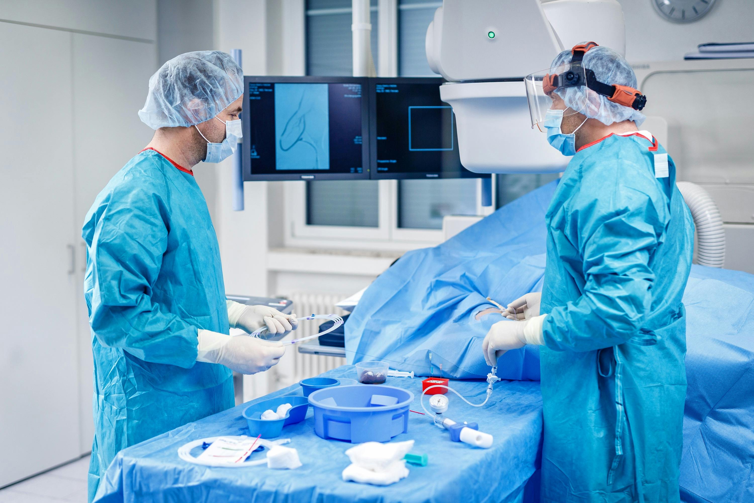 Two surgeons in sterile clothing at work in the operating theatre with medical instruments and a surveillance monitor.