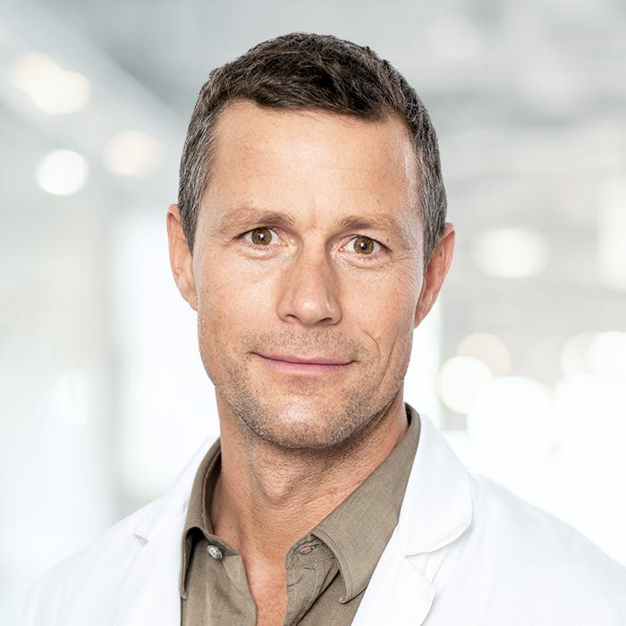 Porträt eines lächelnden Arztes mit weißem Kittel vor unscharfem Hintergrund.