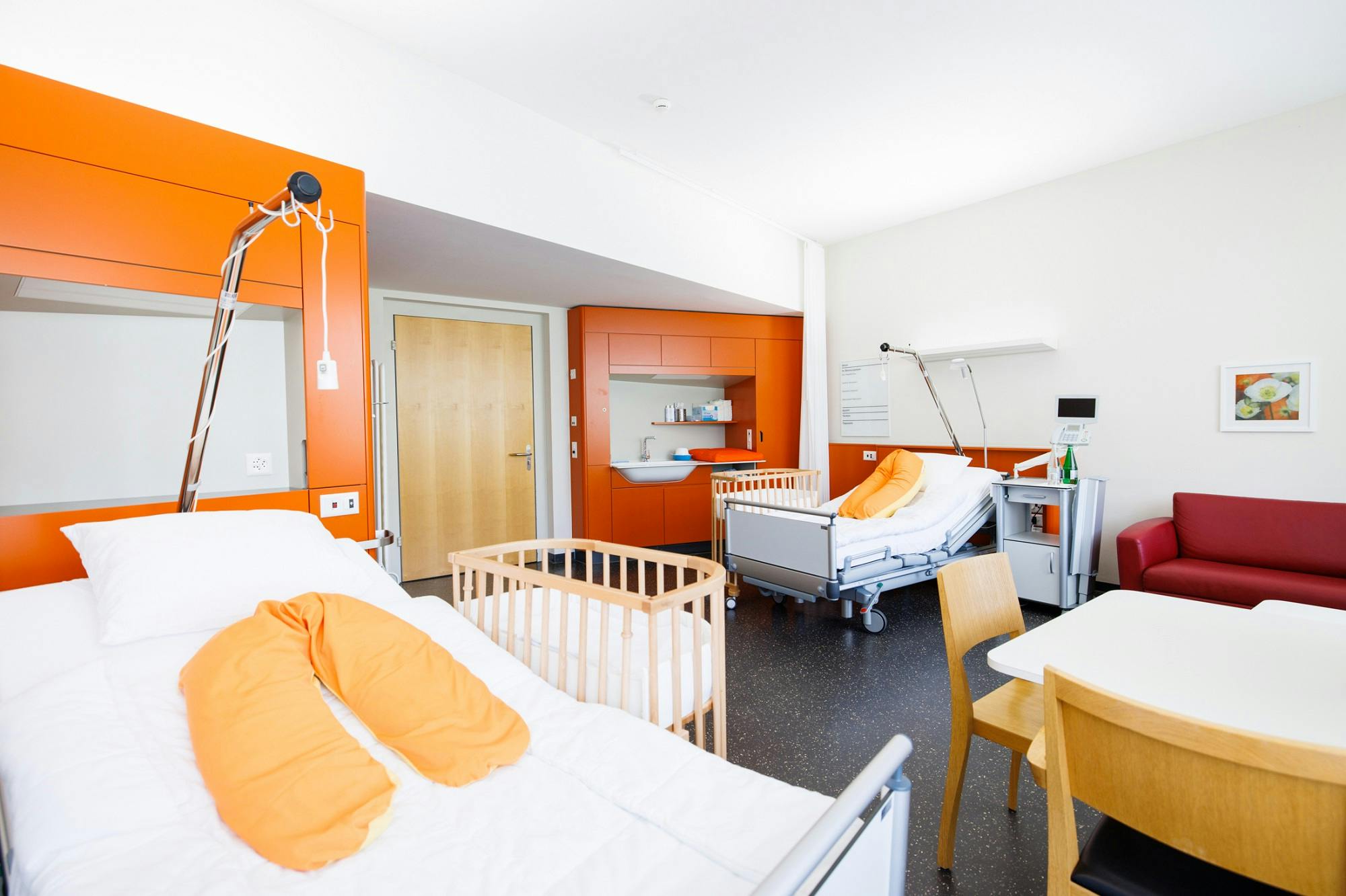Helles Krankenzimmer mit orangefarbenen Akzenten, einem Patientenbett, einem Kinderbett und medizinischer Ausstattung.