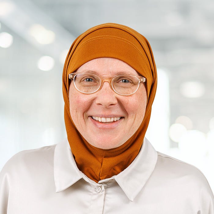 Frau mit Brille und orangefarbenem Hijab lächelt, vor unscharfem Hintergrund.