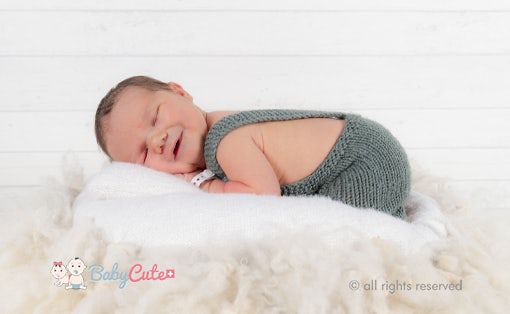 Lächelndes Baby in grünem Strampler liegt auf weißer Decke.