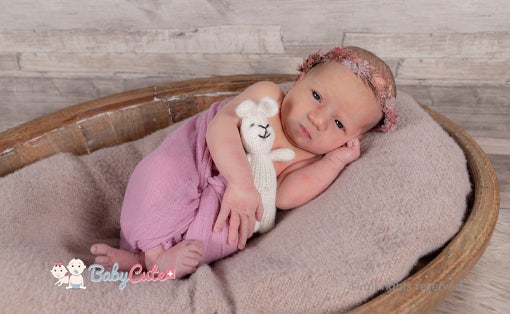 Neugeborenes in rosa Strampler hält ein weißes Stofftier und liegt in einer hölzernen Wanne auf einer braunen Decke.