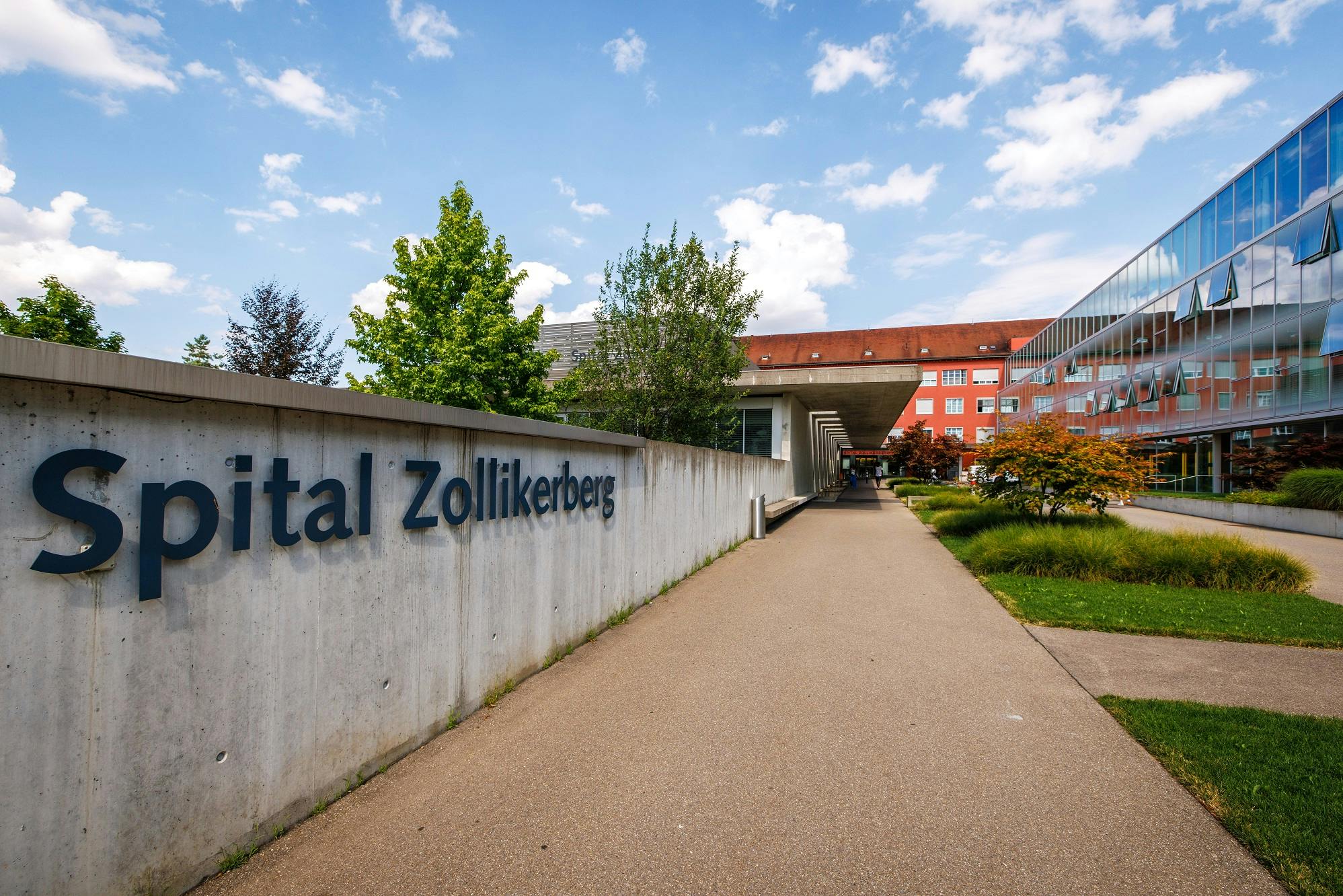 Exterior view of Zollikerberg Hospital with path and green surroundings in sunny weather.
