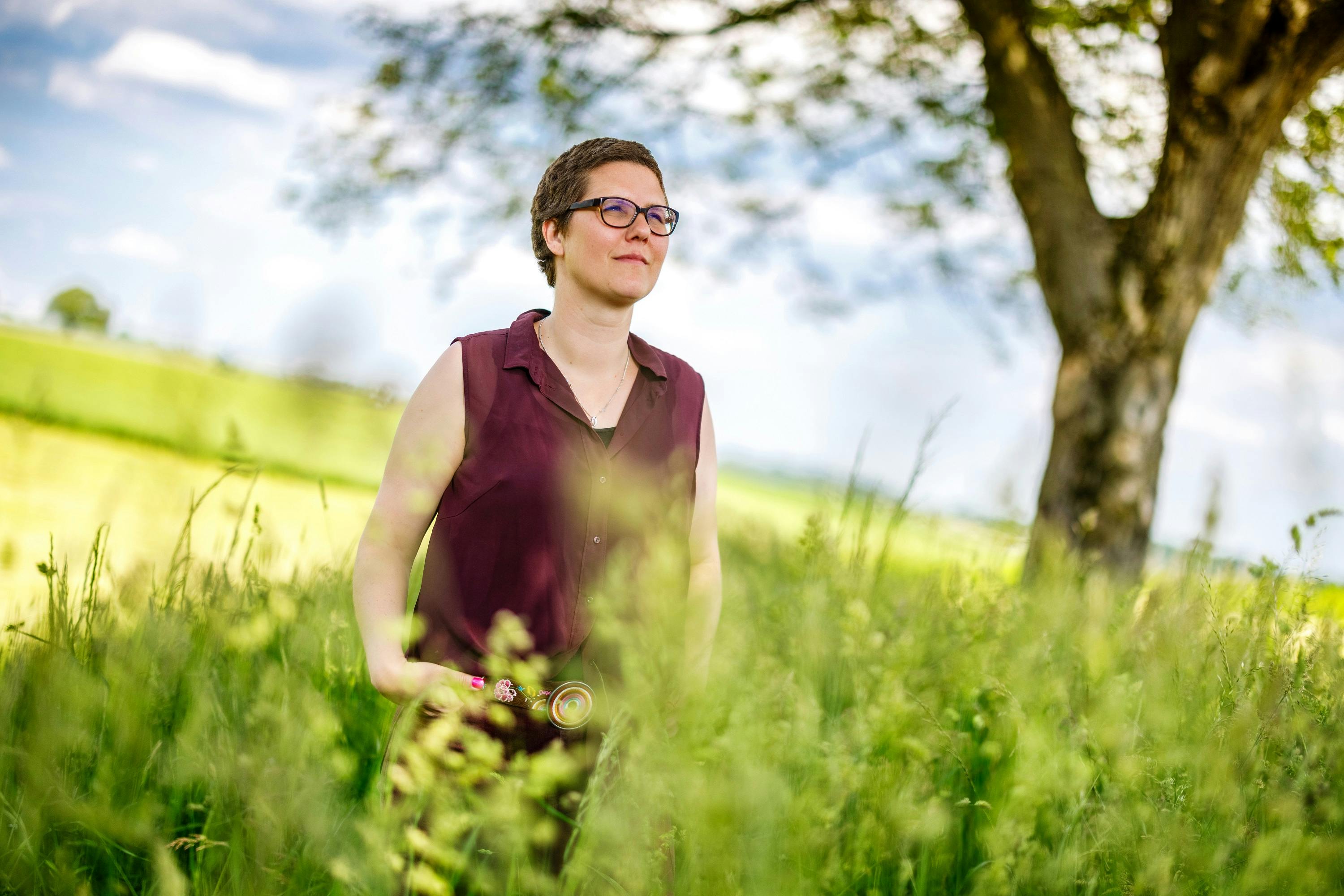 Woman in meadow in sunshine wears glasses and smiles gently.