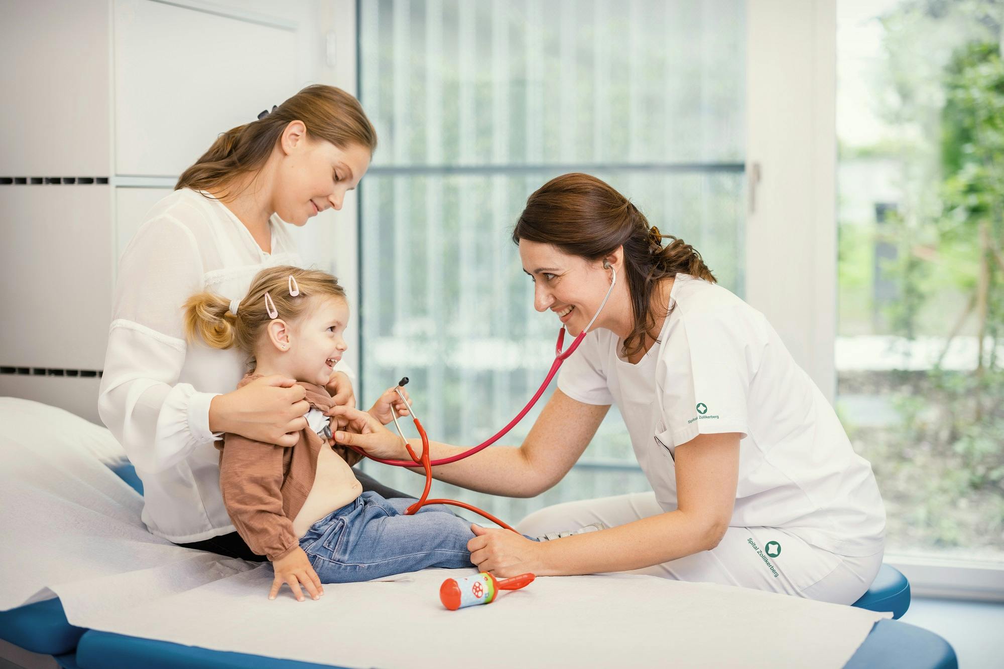 Paediatrician examining smiling girl with stethoscope, mother supporting her.