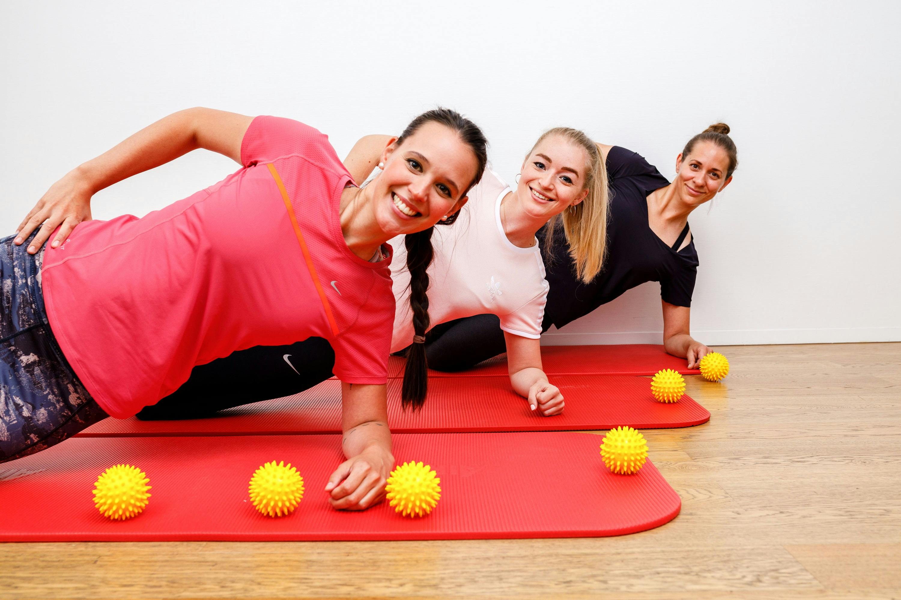 Drei lächelnde Frauen machen Plankenübungen auf roten Fitnessmatten mit gelben Massagebällen.