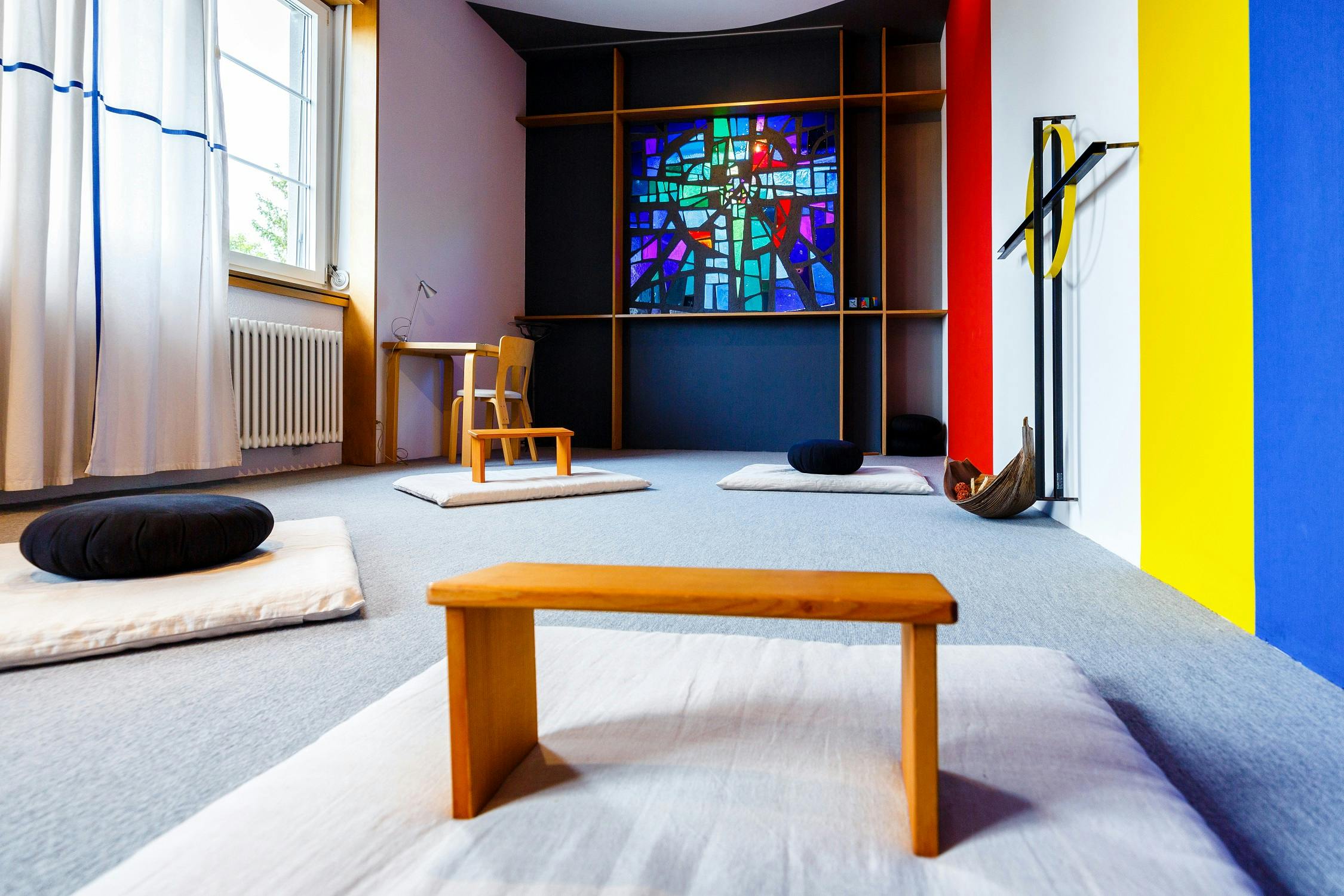 Moderner Meditationsraum mit buntem Kirchenfenster, Kissen und Holzmöbeln.