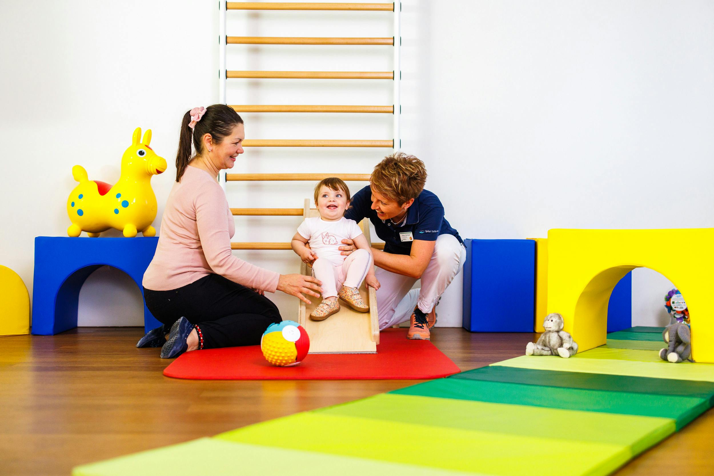 Physiotherapie-Sitzung mit einem Kleinkind und zwei Erwachsenen in einer bunten Praxis.