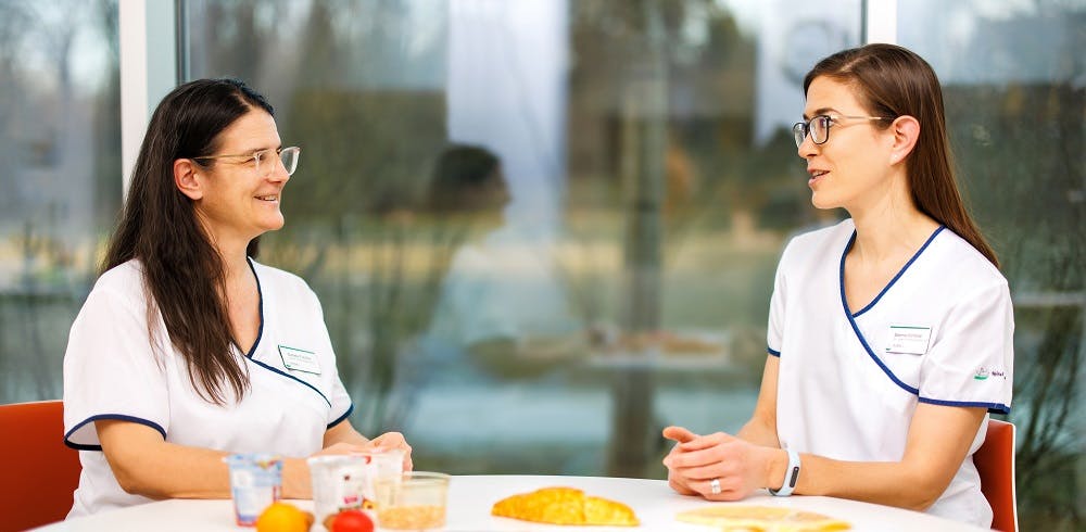 Two people in professional medical clothing talking at a table with food.