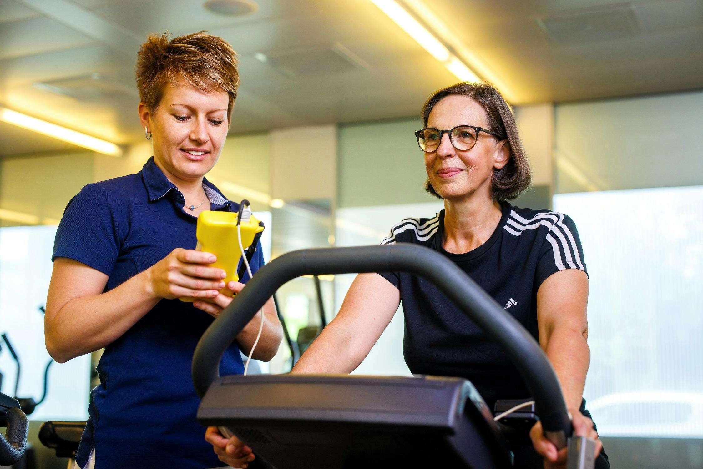 Zwei Frauen im Fitnessstudio, eine beim Training auf dem Laufband und die andere mit einem Smartphone.