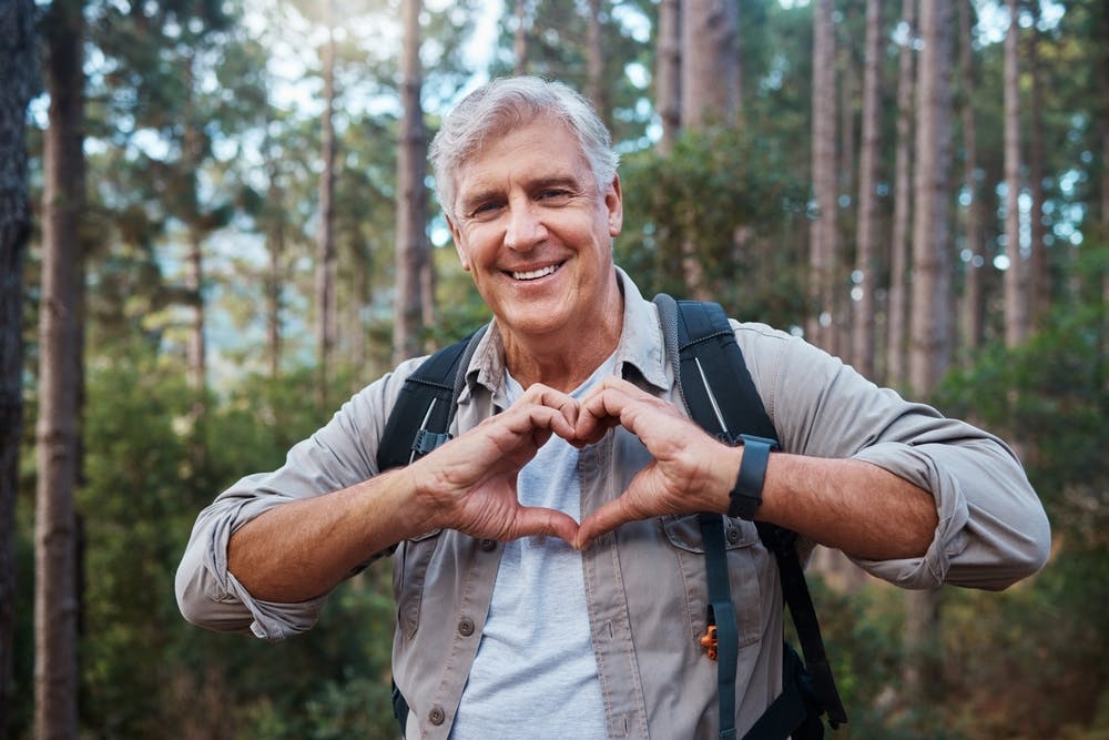 Smiling elderly hiker makes heart gesture in the forest.