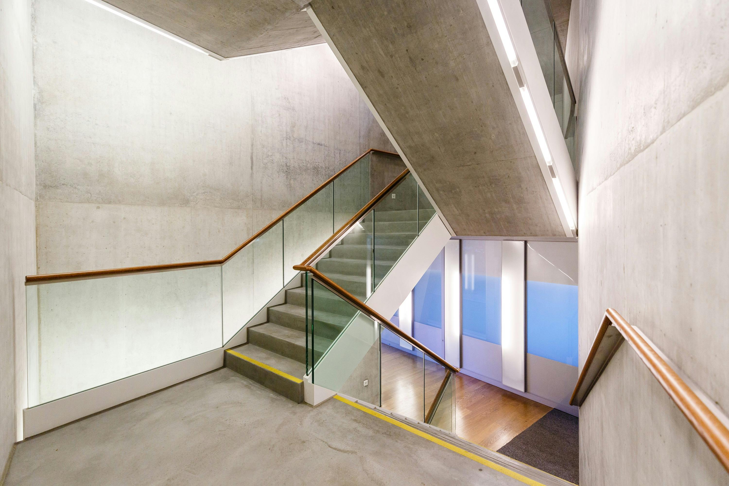 Moderne Betontreppe mit Glasgeländer und holzfarbenem Handlauf in einem zeitgenössischen Gebäude.