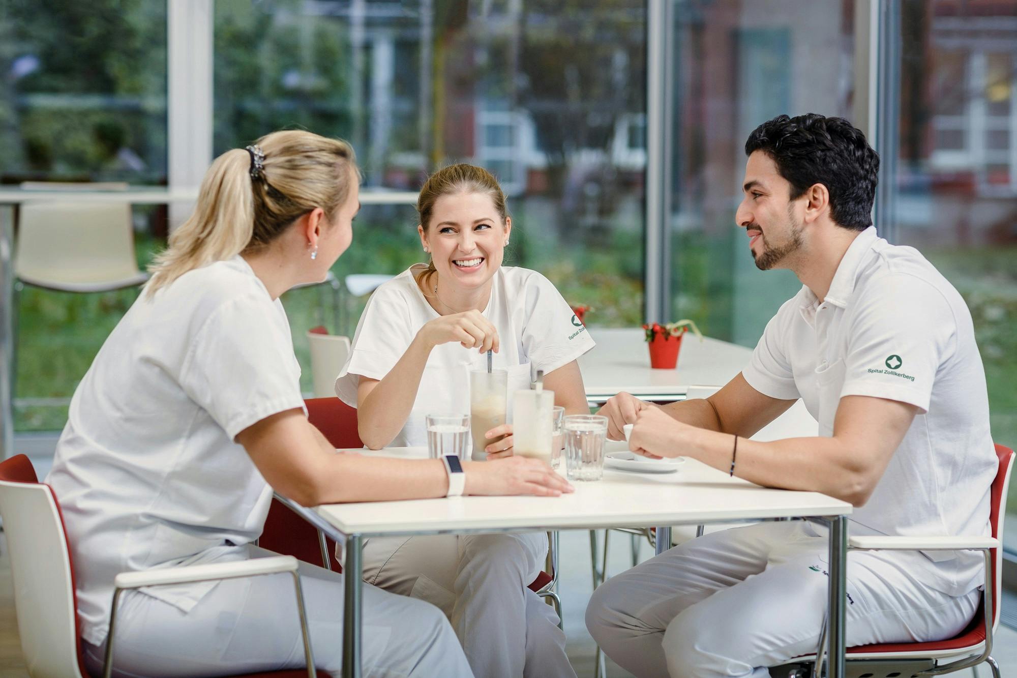 Drei lächelnde Mediziner in weißer Uniform sitzen an einem Tisch im Pausenraum.